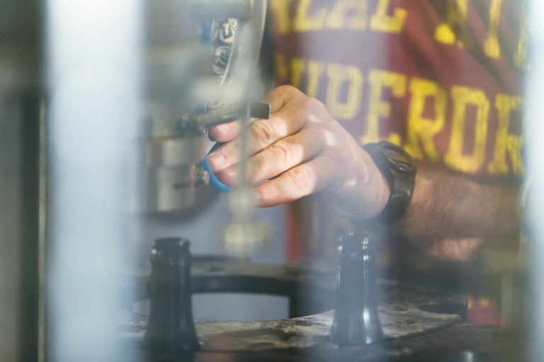 Gros plan sur les mains d'une personne qui manipule une machine dans un environnement industriel. L'individu, vêtu d'une chemise rouge de la Brasserie Gasconha Pessac avec un texte jaune et portant une montre-bracelet noire, ajuste un outil ou un composant sur la machine, entouré d'équipements industriels flous.