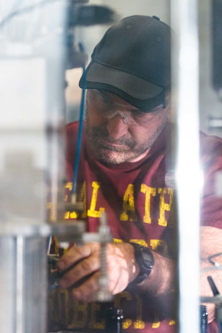 Un homme portant une casquette noire, des lunettes de sécurité et un t-shirt bordeaux avec le texte jaune « Brasserie Gasconha Pessac » travaille intensément sur des machines. Il semble effectuer des réglages ou des réparations, en se concentrant sur une partie spécifique de l'équipement. Son environnement est industriel, suggérant un environnement d'atelier ou d'usine.