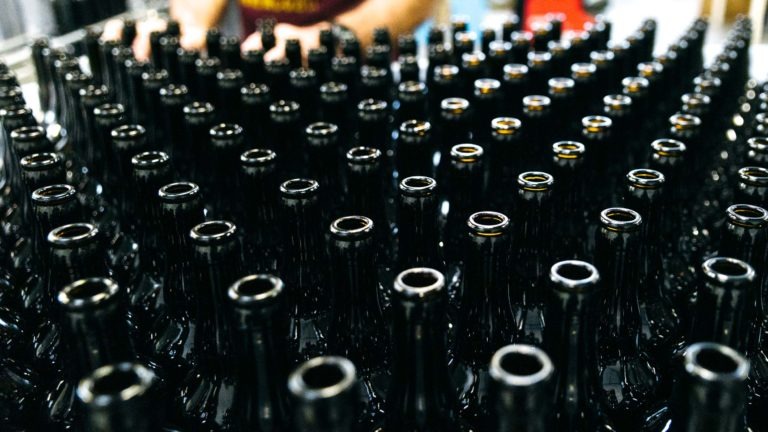 Une grande quantité de bouteilles vides en verre foncé sont serrées les unes contre les autres sur une chaîne de production de la Brasserie Gasconha de Pessac. Les bouteilles sont de taille et d'apparence uniformes, leurs bords brillent sous les lumières du plafond. Dans l'arrière-plan flou, on peut voir une personne en train de mettre en bouteille.