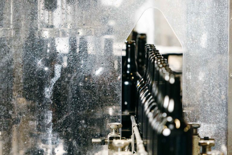 Une rangée de bouteilles en verre noir défile sur un tapis roulant dans l'usine de la Brasserie Gasconha de Pessac. L'image capture le processus de production, montrant les bouteilles passant à travers une section métallique dotée d'un panneau transparent. Le décor industriel met en valeur les machines et le fonctionnement systématique.