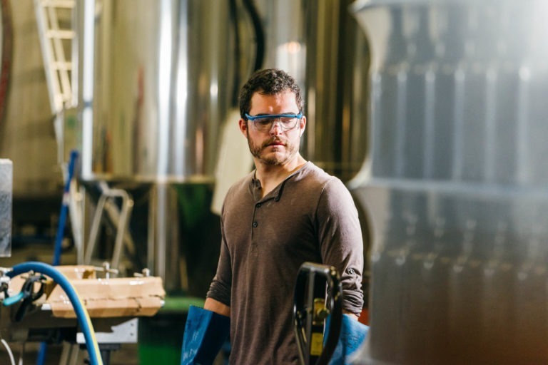 Un homme aux cheveux courts et foncés, barbu, porte une chemise henley marron et des lunettes de sécurité bleues. Il se tient dans un décor industriel avec de grandes cuves métalliques, peut-être la brasserie Gasconha Pessac, en arrière-plan. Il y a un tuyau bleu et une boîte en carton sur le côté gauche de l'image.