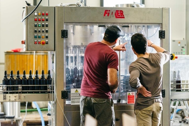 Deux hommes examinent et font fonctionner une machine d'embouteillage FIMER à la Brasserie Gasconha à Pessac. L'homme de gauche montre la machine tandis que celui de droite l'observe. Les deux hommes sont habillés de manière décontractée. Des bouteilles partiellement remplies sont visibles sur la machine, avec d'autres bouteilles vides au premier plan.