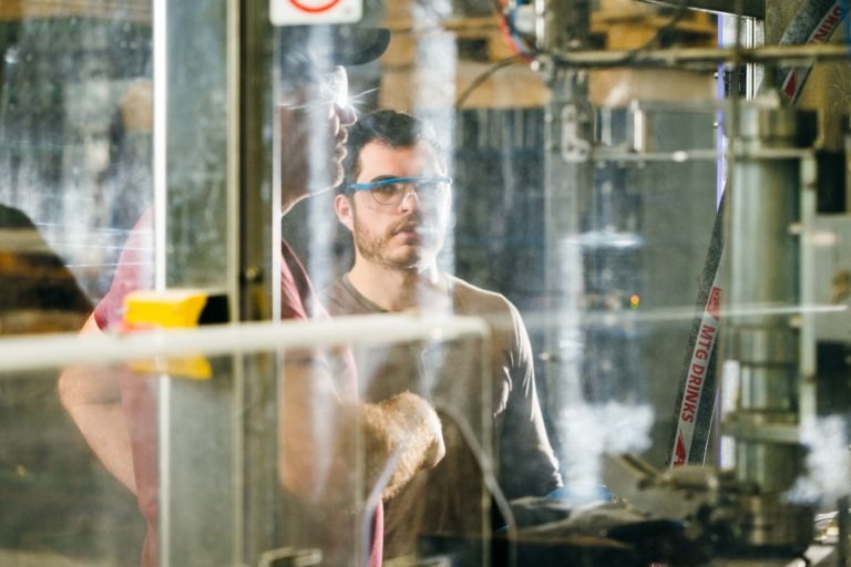 Deux hommes se tiennent dans un environnement semblable à celui d'un atelier à la Brasserie Gasconha de Pessac, concentrés sur les machines. L'un, portant des lunettes de protection et une barbe, observe attentivement, tandis que l'autre, portant une casquette de baseball et des lunettes, explique quelque chose. Le décor est industriel avec des tuyaux, des câbles et divers équipements visibles.