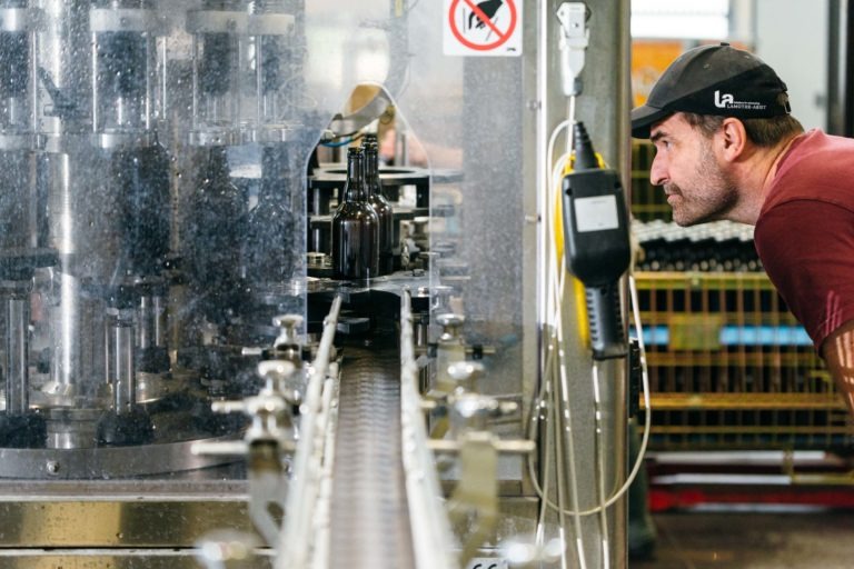 Un homme en chemise rouge et casquette noire inspecte de près des bouteilles qui se déplacent sur un tapis roulant dans une usine. Les bouteilles, provenant probablement de la Brasserie Gasconha Pessac, sont sur une chaîne de production entourée de machines. Un panneau « Interdiction de fumer » est visible en arrière-plan au milieu de divers éléments industriels.