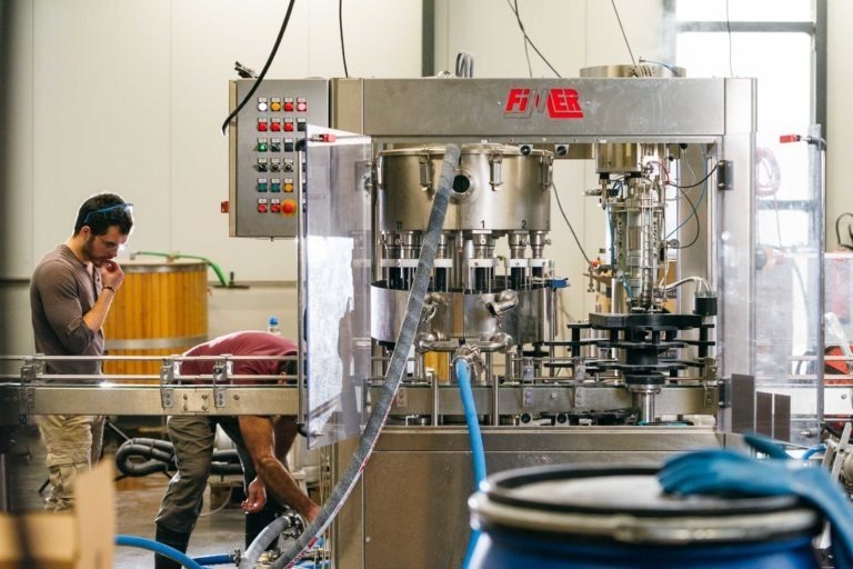 Une machine d'embouteillage fonctionne dans un environnement industriel avec un panneau de commande visible et divers tuyaux et tubes. Deux ouvriers sont présents : l'un travaille activement sur la machine, et l'autre, de la Brasserie Gasconha Pessac, observe avec une expression pensive, la main sur le menton. L'environnement semble industriel.