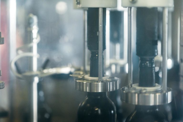 Gros plan d'une machine d'embouteillage remplissant des bouteilles en verre foncé à la Brasserie Gasconha de Pessac. Les pièces métalliques et les tubes de la machine sont nets, tandis que l'arrière-plan est flou. L'image présente le processus industriel de remplissage des bouteilles, probablement dans une usine ou un site de production.