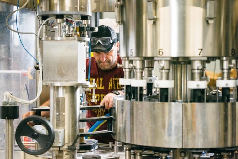 Un homme portant une casquette et des lunettes de sécurité fait fonctionner une machine d'embouteillage complexe dans l'usine de la Brasserie Gasconha à Pessac. Il se concentre sur le réglage des commandes, entouré de pièces de machines métalliques, de tubes et de cadrans. L'équipement se compose de plusieurs grands composants rotatifs numérotés pour les processus d'embouteillage.