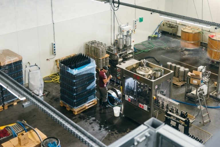 Une personne fait fonctionner des machines dans la Brasserie Gasconha Pessac, debout à côté d'un panneau de commande avec divers boutons et écrans. Autour des machines se trouvent des palettes de bouteilles empilées, des cuves métalliques reliées par des tuyaux et d'autres équipements de brassage. Le sol est humide et l'espace semble être une zone de production.