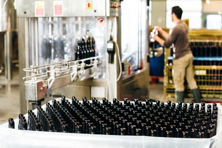 Une chaîne de production de la Brasserie Gasconha de Pessac présente de nombreuses bouteilles en verre brun vides alignées dans un récipient en plastique en attente d'être remplies. À l'arrière-plan, une personne aux cheveux noirs et aux vêtements gris fait fonctionner une machine qui place des bouteilles sur un tapis roulant. Des piles de cartons sont visibles au loin.