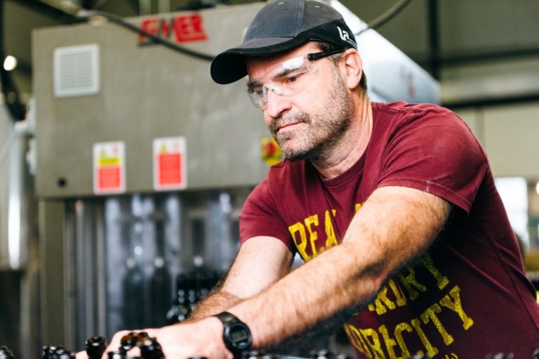 Un homme portant des lunettes de sécurité et une casquette noire fait fonctionner des machines dans un environnement industriel, probablement à la Brasserie Gasconha de Pessac. Il est vêtu d'un T-shirt marron avec un texte jaune et d'une montre-bracelet noire. En arrière-plan, des équipements métalliques et des bouteilles s'alignent sur un tapis roulant, suggérant qu'il se trouve dans une usine ou un atelier.