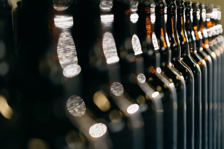 Gros plan d'une rangée de bouteilles en verre foncé de la Brasserie Gasconha Pessac, avec des lumières floues se reflétant sur leurs surfaces. Les bouteilles sont alignées avec soin, s'étendant au loin et créant un motif répétitif. Les reflets de lumière créent des effets bokeh doux sur la texture brillante des bouteilles.