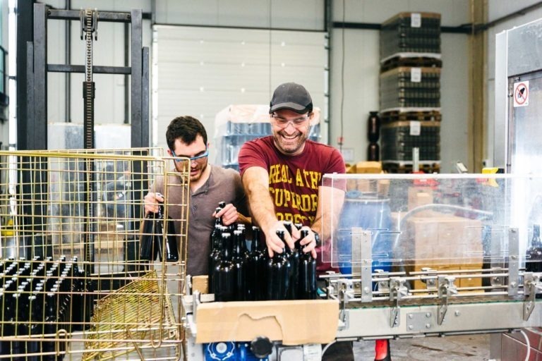 Deux hommes travaillent dans l'usine d'embouteillage de la Brasserie Gasconha Pessac. L'homme de droite, portant une casquette et un t-shirt rouge, sourit tout en manipulant des bouteilles en verre foncé sur un convoyeur industriel. L'homme de gauche, portant des lunettes, ajuste des équipements. Ils sont entourés de machines et d'étagères sur lesquelles sont empilées des palettes.