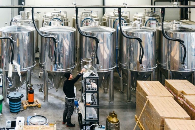 Un ouvrier de la brasserie Gasconha à Pessac vérifie l'une des nombreuses cuves de fermentation en acier inoxydable. La pièce est remplie d'équipements de brassage, notamment de tuyaux et de flexibles reliés aux cuves. À proximité, des cartons et divers conteneurs sont empilés sur des palettes et des étagères.