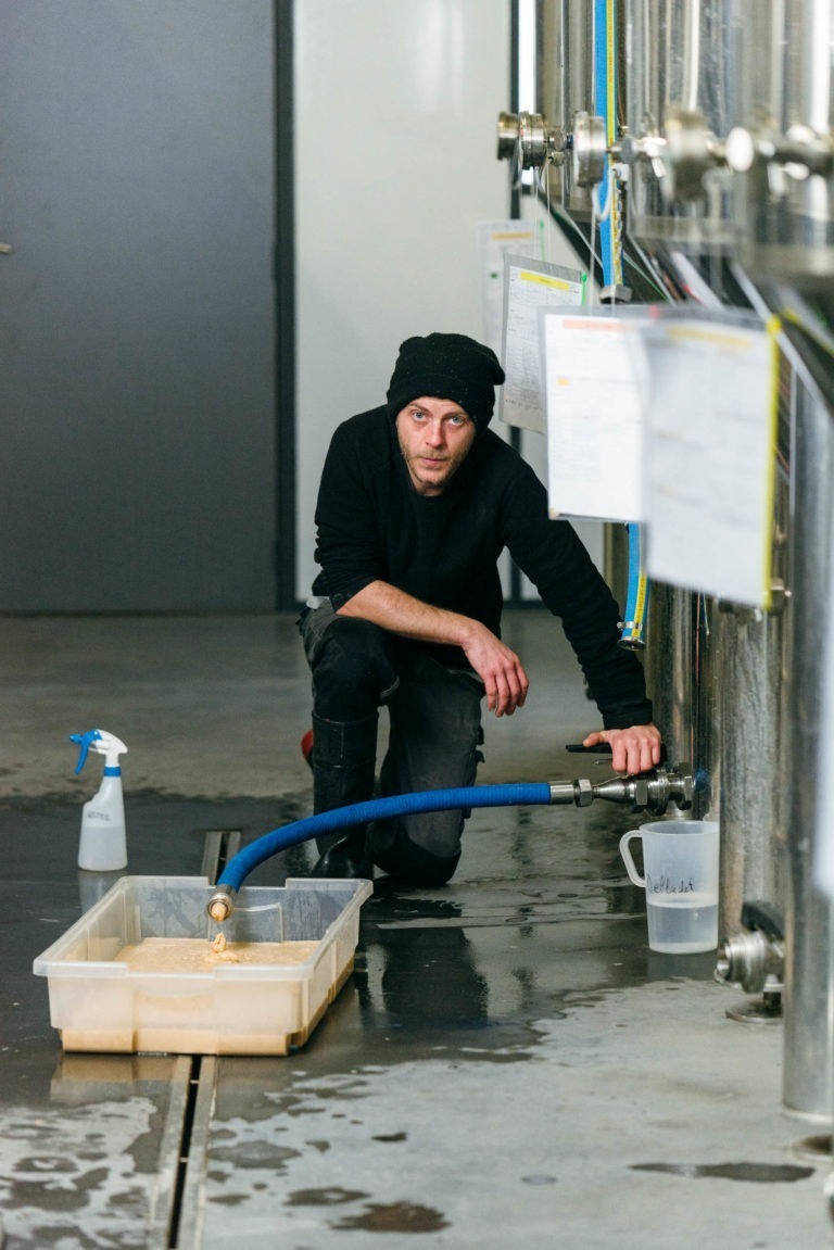 Une personne est agenouillée à côté d'un grand réservoir en métal avec divers tuyaux et panneaux de contrôle à la Brasserie Gasconha de Pessac. Elle tient une valve attachée à un tuyau bleu, remplissant un récipient en plastique transparent de liquide. Un vaporisateur est posé sur le sol mouillé à proximité. La personne porte des vêtements noirs et un bonnet noir.