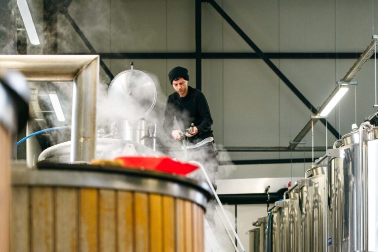 Une personne portant un bonnet noir et des vêtements sombres se tient debout sur un escabeau tout en travaillant avec une grande cuve de brassage en acier inoxydable fumante à la Brasserie Gasconha Pessac. L'installation dispose d'équipements de brassage industriels avec plusieurs cuves métalliques disposées en arrière-plan. Le décor semble être celui d'une brasserie.