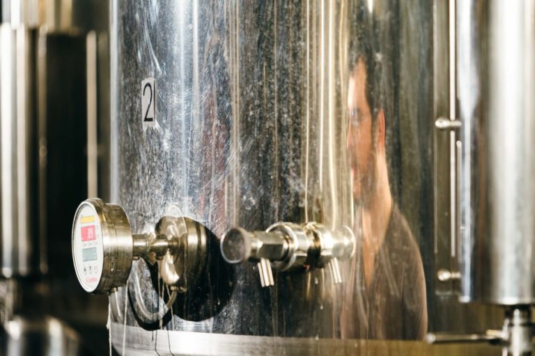 Une cuve de fermentation métallique portant le numéro « 2 » est présentée à la Brasserie Gasconha Pessac. La cuve est dotée de divers tuyaux et d'une jauge affichant « 1,5 » en rouge et blanc. Le reflet d'un homme portant des lunettes est visible sur la surface brillante alors qu'il semble inspecter la cuve.