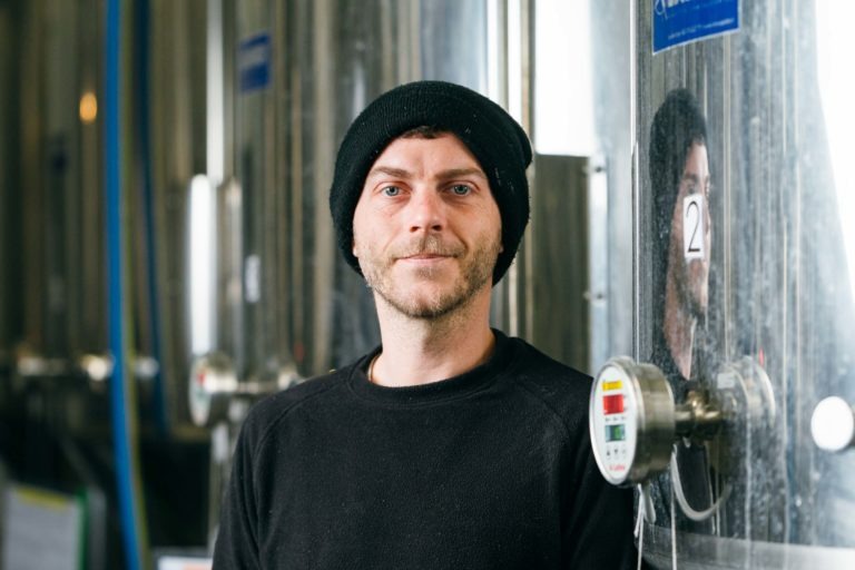 Un homme portant un bonnet et un pull noirs se tient devant des cuves industrielles en acier inoxydable à la Brasserie Gasconha de Pessac. Son reflet scintille sur l'une des cuves. L'arrière-plan comprend des surfaces métalliques, des tuyaux et une jauge partiellement visible, soulignant le cadre professionnel et industriel.