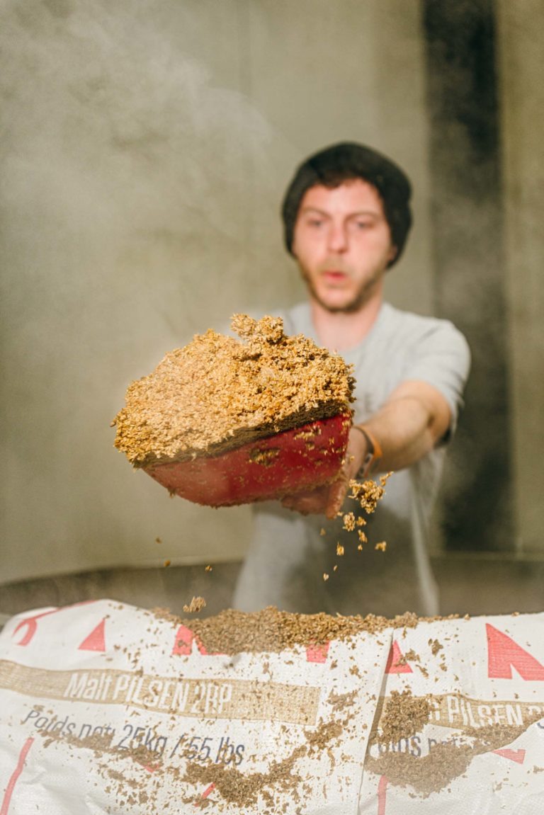 Un homme portant un bonnet et une chemise grise tient une pelle rouge remplie de malt au-dessus d'un grand récipient ou d'un sac étiqueté « Malt PILSEN ». L'arrière-plan flou suggère le brassage à la Brasserie Gasconha à Pessac. Concentré sur sa tâche, l'individu incarne le savoir-faire et le dévouement du processus de brassage.