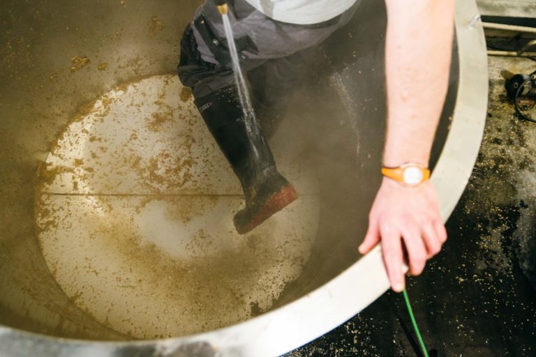 Une personne nettoie l'intérieur d'un grand récipient en métal avec un jet d'eau à haute pression à la Brasserie Gasconha de Pessac. Le bas du corps et les jambes, chaussés de bottes en caoutchouc noires, sont visibles. La main droite de la personne, ornée d'une montre-bracelet, repose sur le bord du récipient tandis que de la vapeur et des gouttes d'eau remplissent l'espace.