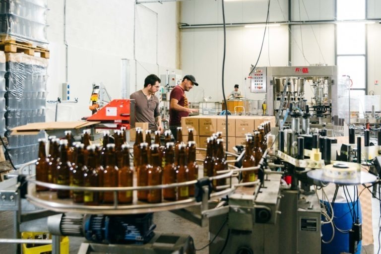 Deux hommes travaillent dans la brasserie Gasconha de Pessac. L'un place des caisses sur une table, tandis que l'autre fait fonctionner des machines. Au premier plan, des bouteilles en verre brun sont disposées sur un tapis roulant, prêtes à être remplies. La pièce est bien éclairée, avec des machines industrielles et des caisses empilées en arrière-plan.