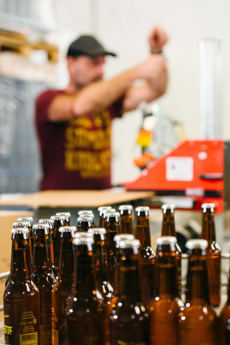 Au premier plan, une rangée de bouteilles de bière en verre ambré avec des capsules en métal est posée sur un tapis roulant. À l'arrière-plan flou, un homme portant une chemise marron et une casquette noire ajuste le matériel de la brasserie Gasconha Pessac. Une machine orange est partiellement visible à côté de lui.