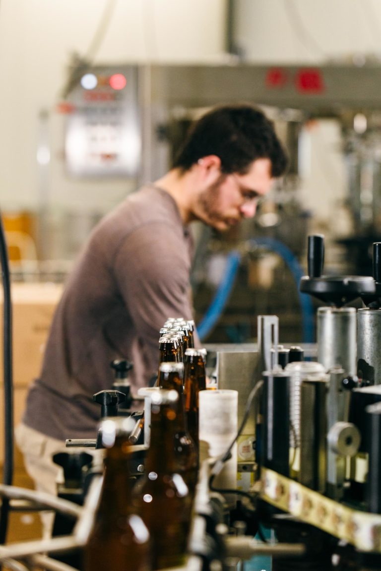 Un homme aux cheveux noirs courts et à la barbe travaille dans la brasserie Gasconha de Pessac, vêtu d'une chemise marron à manches longues et de lunettes de sécurité. Il est concentré sur les tâches liées aux machines de brassage. Au premier plan, plusieurs bouteilles de bière brune sont positionnées sur un système de tapis roulant. L'arrière-plan est légèrement flou.