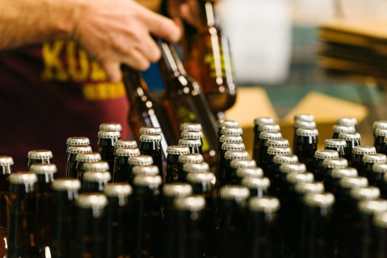 Gros plan d'une personne tenant plusieurs bouteilles de bière de couleur ambre de la Brasserie Gasconha Pessac avec une main au-dessus d'un grand groupe de bouteilles de bière bouchées sur un tapis roulant. La personne porte un t-shirt avec un texte partiel et indiscernable. L'arrière-plan est flou, ce qui concentre l'attention sur les bouteilles.