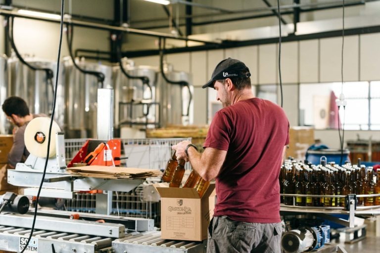 À la Brasserie Gasconha de Pessac, un homme en chemise rouge et casquette noire emballe des boissons en bouteille dans une boîte en carton sur la chaîne de production. Le tapis roulant déplace les bouteilles et les matériaux d'emballage, tandis que des équipements industriels et un espace de travail bien éclairé avec de grandes cuves métalliques sont visibles en arrière-plan.