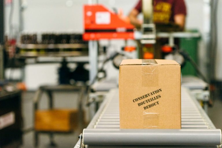 Une boîte en carton étiquetée "CONSERVATION BOUTEILLES DEBOUT" est posée sur un tapis roulant dans l'usine de la Brasserie Gasconha à Pessac. L'arrière-plan montre des machines floues et une personne en chemise marron, légèrement floue. La boîte est fermée avec du ruban adhésif et positionnée au centre de l'image.