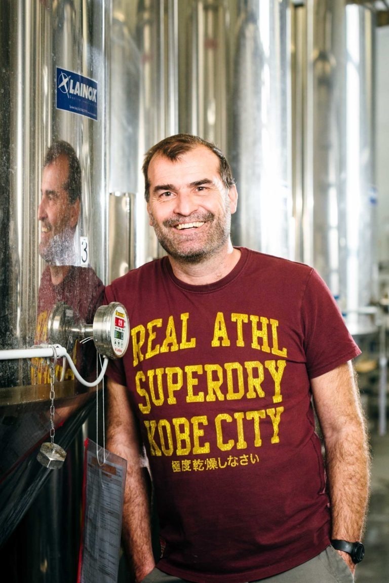 Un homme barbu aux cheveux courts sourit debout dans un décor industriel, peut-être une brasserie comme la Brasserie Gasconha Pessac, à côté d'une grande cuve en acier inoxydable. Il porte un T-shirt marron avec le texte jaune "REAL ATHL SUPERDRY KOBE CITY". Son reflet peut être vu dans la surface brillante de la cuve.