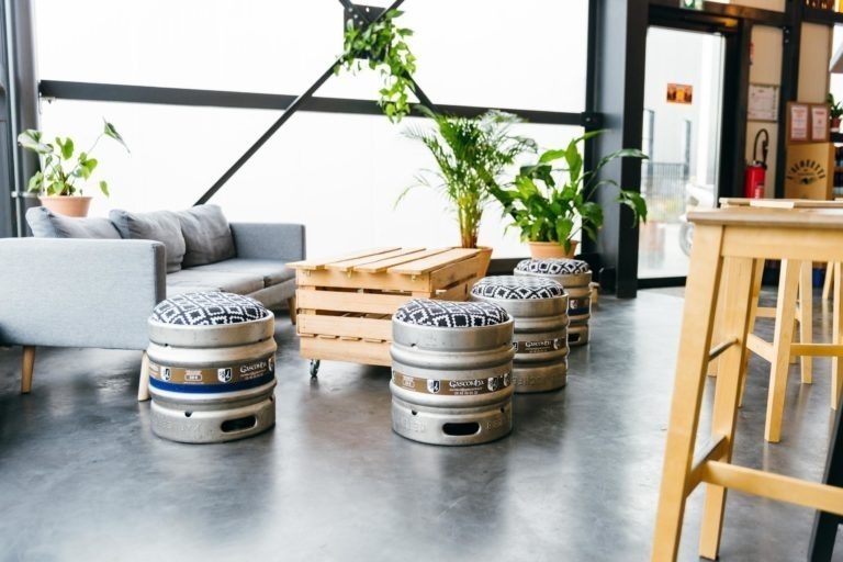 Espace salon intérieur moderne avec un canapé gris à gauche, une table basse en palette de bois au centre et quatre tabourets fabriqués à partir de fûts de bière en métal avec des coussins à motifs. L'espace est orné de plantes vertes en pot, de grandes fenêtres en verre et d'un tabouret de bar de la Brasserie Gasconha Pessac visible sur le côté droit.