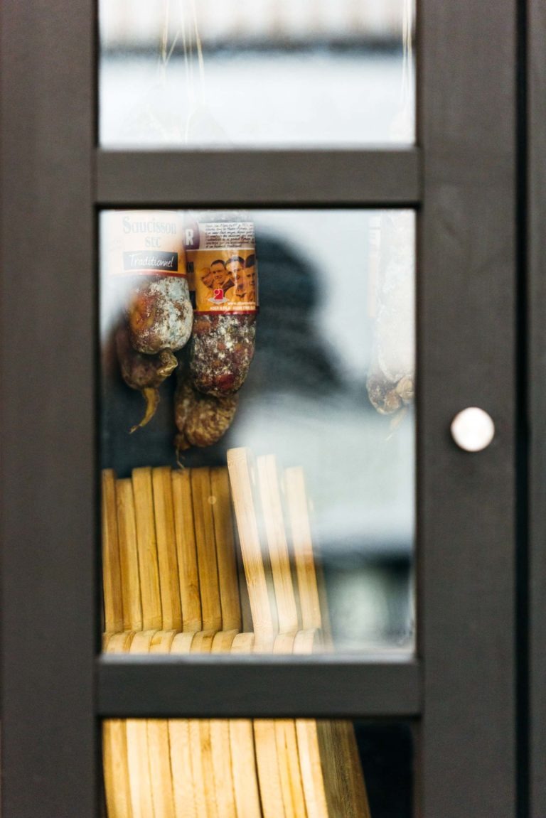 Une vue rapprochée à travers une porte vitrée révèle des saucisses séchées suspendues et des planches de bois soigneusement empilées à l'intérieur de la Brasserie Gasconha Pessac. Les saucisses sont emballées avec des étiquettes aux motifs détaillés. Le cadre de la porte est sombre avec une poignée métallique ronde sur le côté droit.