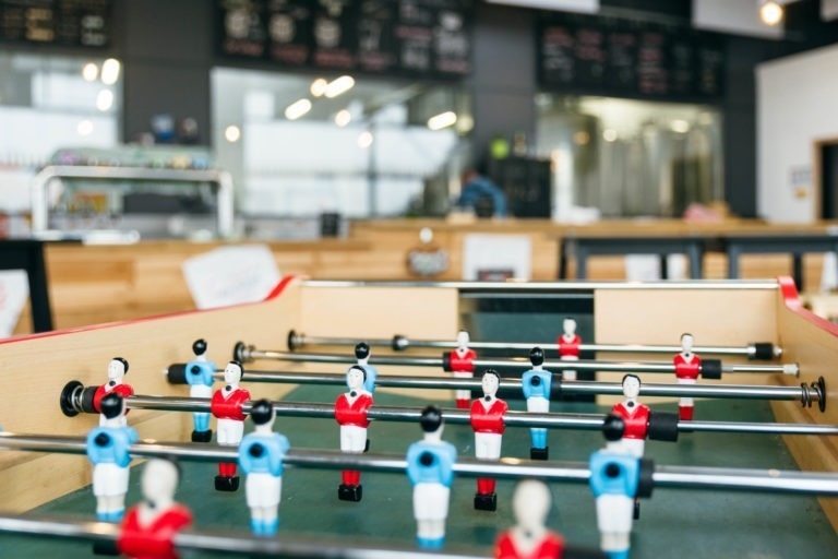 Gros plan d'un baby-foot avec des figurines de joueurs rouges et bleues positionnées comme si elles étaient en train de jouer. La scène se déroule dans une salle très éclairée de la Brasserie Gasconha Pessac, avec un arrière-plan flou comportant des comptoirs en bois, des tabourets de bar et un panneau de menu, suggérant que le décor pourrait être un café ou un bar.