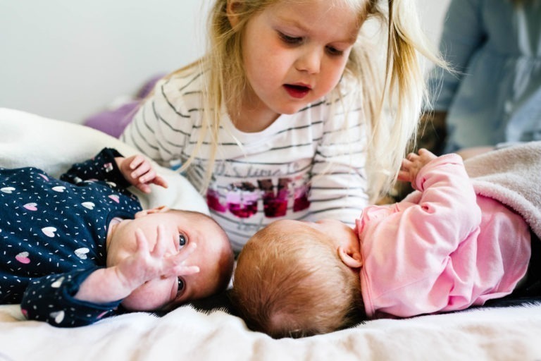 Une jeune fille aux cheveux blonds se penche vers deux bébés couchés côte à côte sur un lit, vêtus d'une chemise blanche à rayures. L'un des bébés est habillé d'une combinaison bleu marine à motifs de cœurs, tandis que l'autre bébé porte une tenue rose. Ils sont tous engagés et se regardent, partageant un moment innocent de coup de foudre au feu rouge.