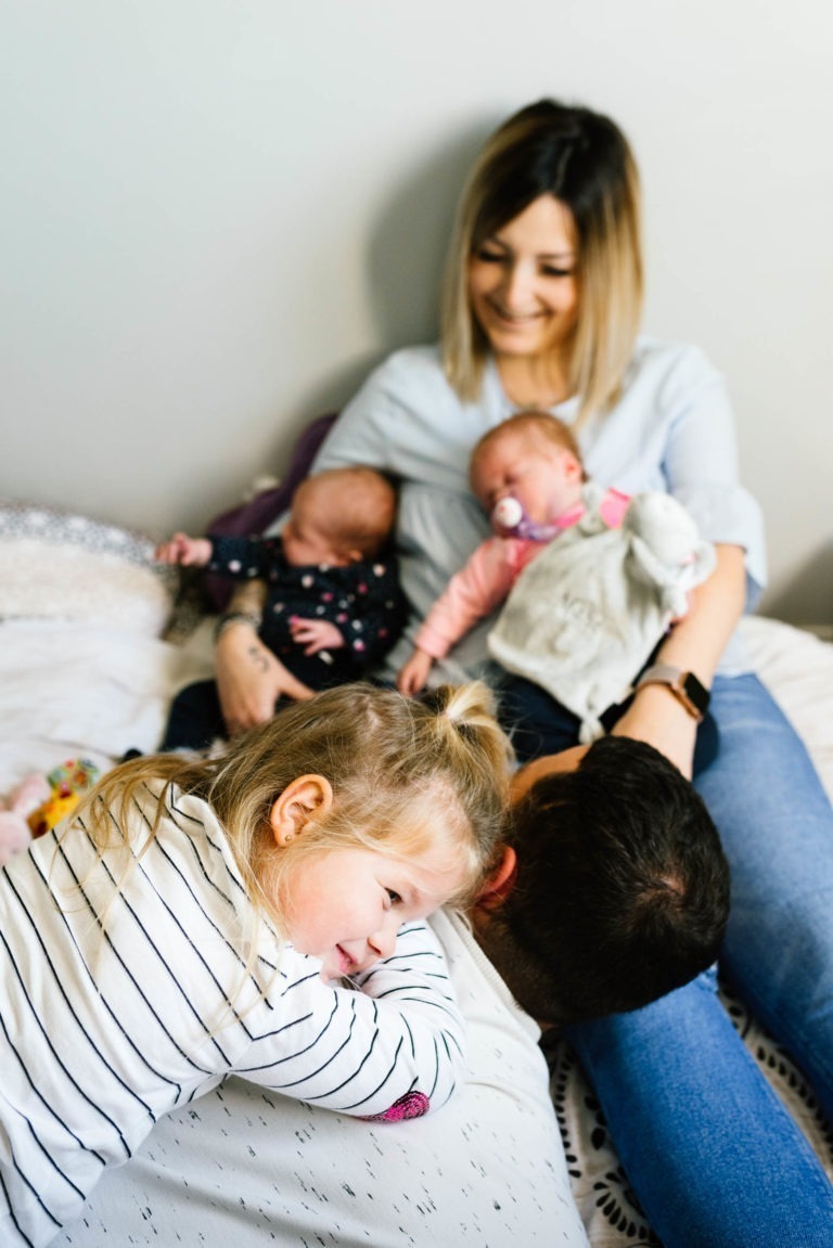 Une mère est assise sur un lit, tenant dans ses bras des jumeaux nouveau-nés. Le bébé de gauche est habillé de noir, tandis que celui de droite porte une tétine et une tenue blanche. Une jeune fille blonde, vêtue d’une chemise rayée, est allongée sur le dos de son père, souriant à la caméra – coup de foudre au feu rouge.