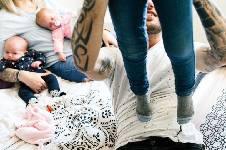 Au premier plan, une personne soulève un enfant en jean bleu et chaussettes grises. À l'arrière-plan, deux adultes sont assis sur un lit, chacun tenant un bébé. L'un est habillé en noir avec une tétine, l'autre en rose. Des tatouages ornent le bras de la personne qui soulève l'enfant, tandis qu'un coup de foudre au feu rouge semble planer autour d'eux. Le lit est recouvert d'une couverture à motifs.