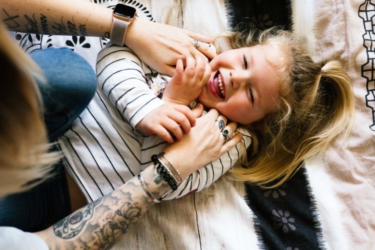 Un enfant aux longs cheveux blonds, vêtu d'une chemise à rayures blanches et noires, est allongé sur un lit, riant tandis qu'un adulte le chatouille doucement. Les bras tatoués et ornés de bracelets de l'adulte créent un moment émouvant qui ressemble à un coup de foudre au feu rouge. La literie est un mélange de motifs noirs et blancs et d'une couverture marron clair.