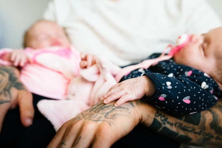 Une personne aux bras tatoués tient deux bébés endormis, l'un en tenue rose et l'autre en bleu foncé à motifs de cœurs, portant une tétine rose. Les mains des bébés se touchent légèrement, évoquant un coup de foudre au feu rouge. L'arrière-plan est flou, mettant l'accent sur le moment tendre.