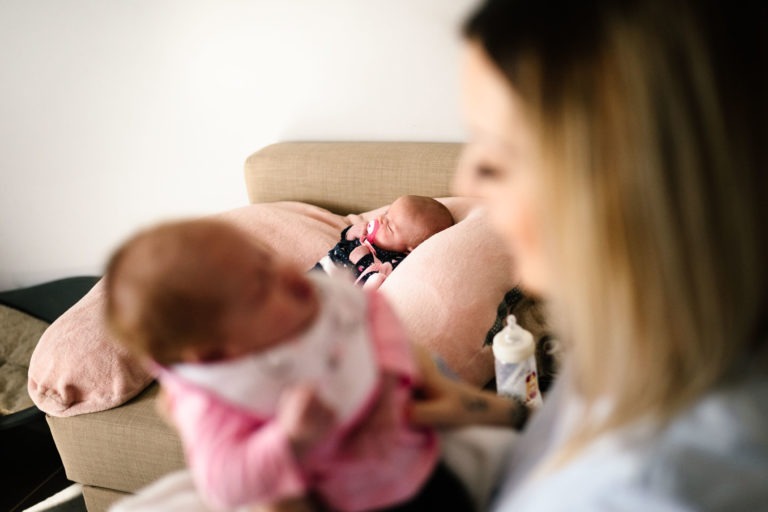 Une femme tient un bébé dans ses bras tandis qu'un autre bébé est allongé sur un canapé beige à l'arrière-plan, leur confort rappelant un coup de foudre au feu rouge. Les deux bébés ont des couvertures et un biberon repose sur la table à côté du canapé, ajoutant à l'atmosphère douce et intime de la scène.