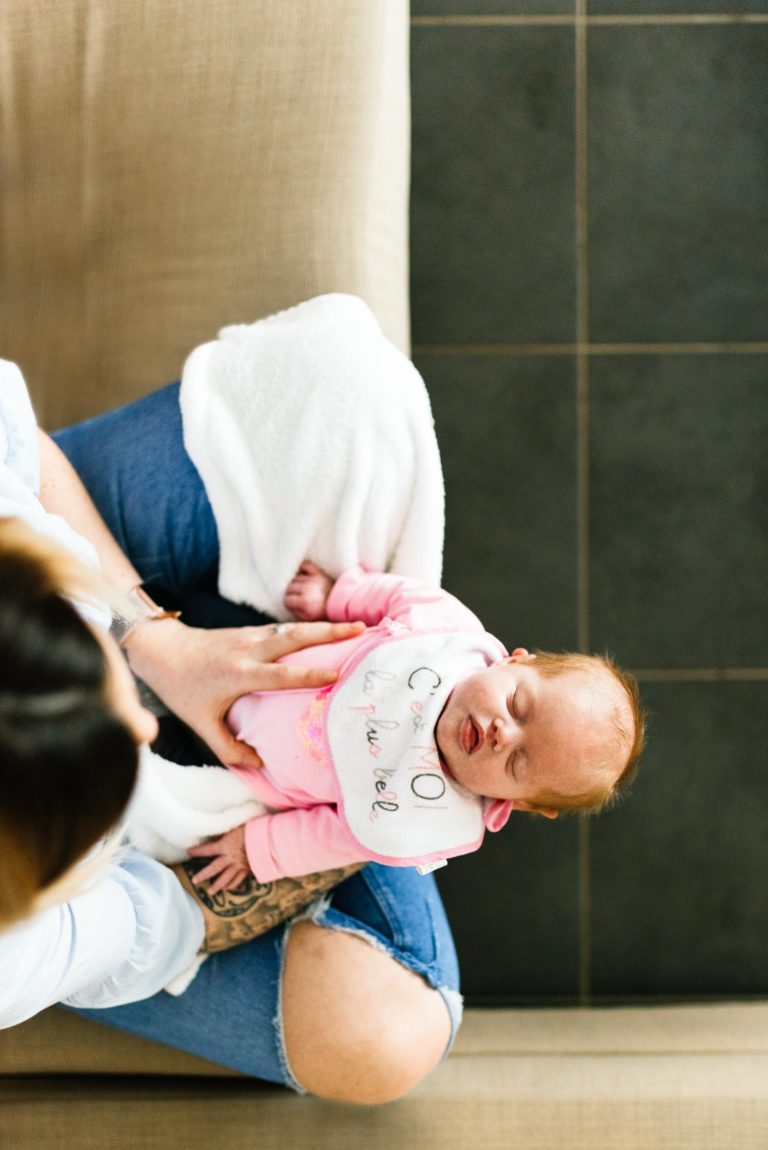 Un bébé habillé en rose, portant un bavoir sur lequel est écrit « Chillin' is my cardio » (se détendre, c'est mon cardio), est allongé sur les genoux d'un adulte assis sur un canapé beige. L'adulte, vêtu d'une chemise blanche et d'un jean bleu, tient doucement le bébé sous une couverture blanche. C'est comme s'ils avaient vécu un coup de foudre au feu rouge, un coup de foudre instantané.