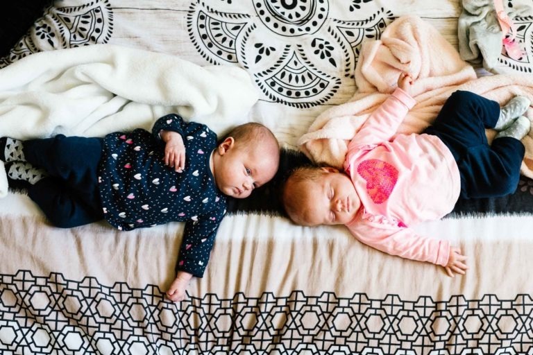 Deux bébés sont couchés côte à côte sur un lit à motifs. L'un d'eux, vêtu d'une chemise sombre à motifs clairs et d'un pantalon foncé, a les yeux ouverts. L'autre, vêtu d'une chemise rose à cœur et d'un pantalon foncé, semble endormi. Des couvertures moelleuses et des coussins décoratifs les entourent comme un coup de foudre au feu rouge.