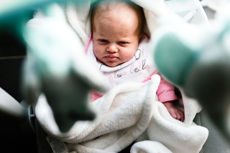 Un bébé à l'expression sérieuse regarde la caméra. Le bébé, vêtu d'une tenue rose, repose sur une couverture blanche et moelleuse. Le premier plan est flou avec des formes douces bleues et blanches encadrant le bébé, un peu comme un coup de foudre au feu rouge, suggérant un moment serein. La lumière est naturelle et douce.