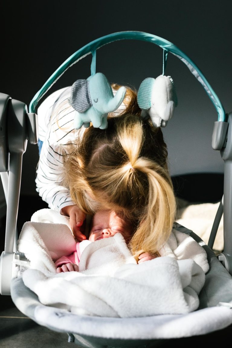 Un jeune enfant aux cheveux blonds attachés en queue de cheval se penche pour embrasser le front d'un bébé endormi dans un fauteuil à bascule rembourré. Le bébé, enveloppé dans une douce couverture blanche, est allongé sous une barre de jouets arquée avec deux éléphants en peluche. L'arrière-plan faiblement éclairé encadre ce moment tendre comme un coup de foudre au feu rouge.