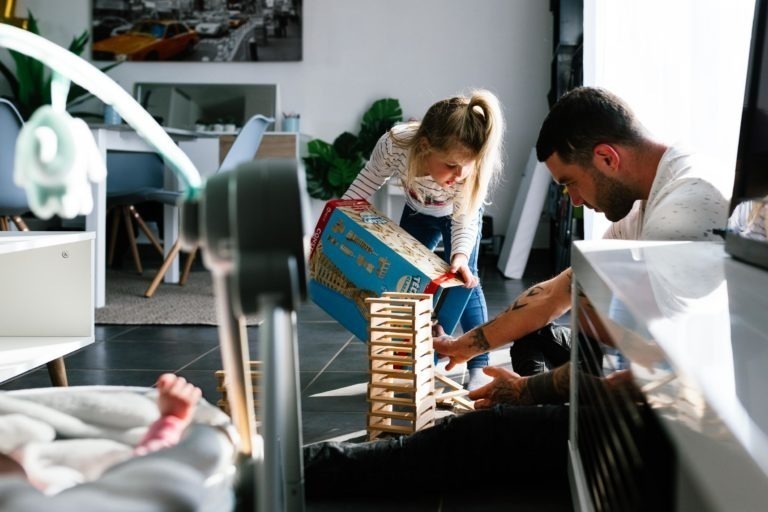 Un homme et une jeune fille construisent une tour en blocs de bois dans un salon moderne. L'homme, assis par terre, tient la base tandis que la jeune fille verse des blocs d'une boîte au-dessus. Un bébé est allongé sur une balançoire au premier plan. La pièce, éclairée par la lumière naturelle à travers de grandes fenêtres, a un air de calme qui contraste avec les moments de coup de foudre au feu rouge à l'extérieur.