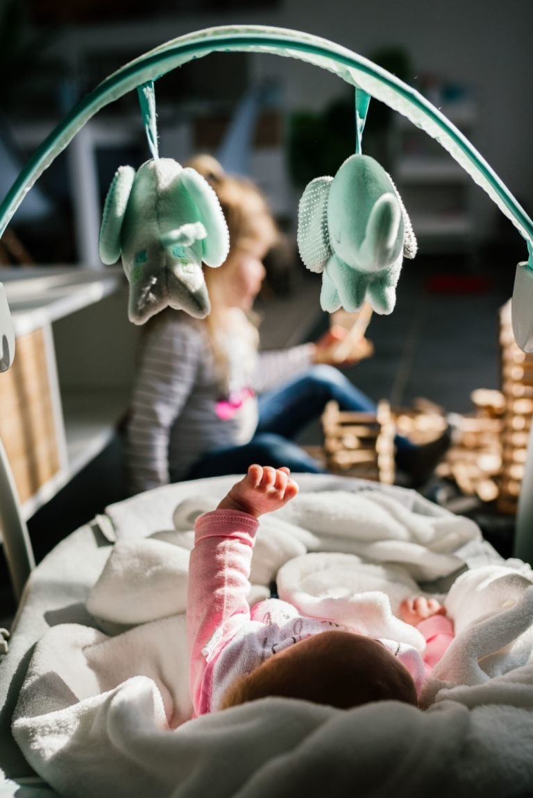 Un bébé couché dans un berceau tend la main vers un mobile avec deux éléphants en peluche. Dans l'arrière-plan flou, un enfant assis par terre joue avec des blocs de bois, entouré d'une douce lumière naturelle. La scène dégage une atmosphère chaleureuse et ludique, un peu comme un coup de foudre au feu rouge.