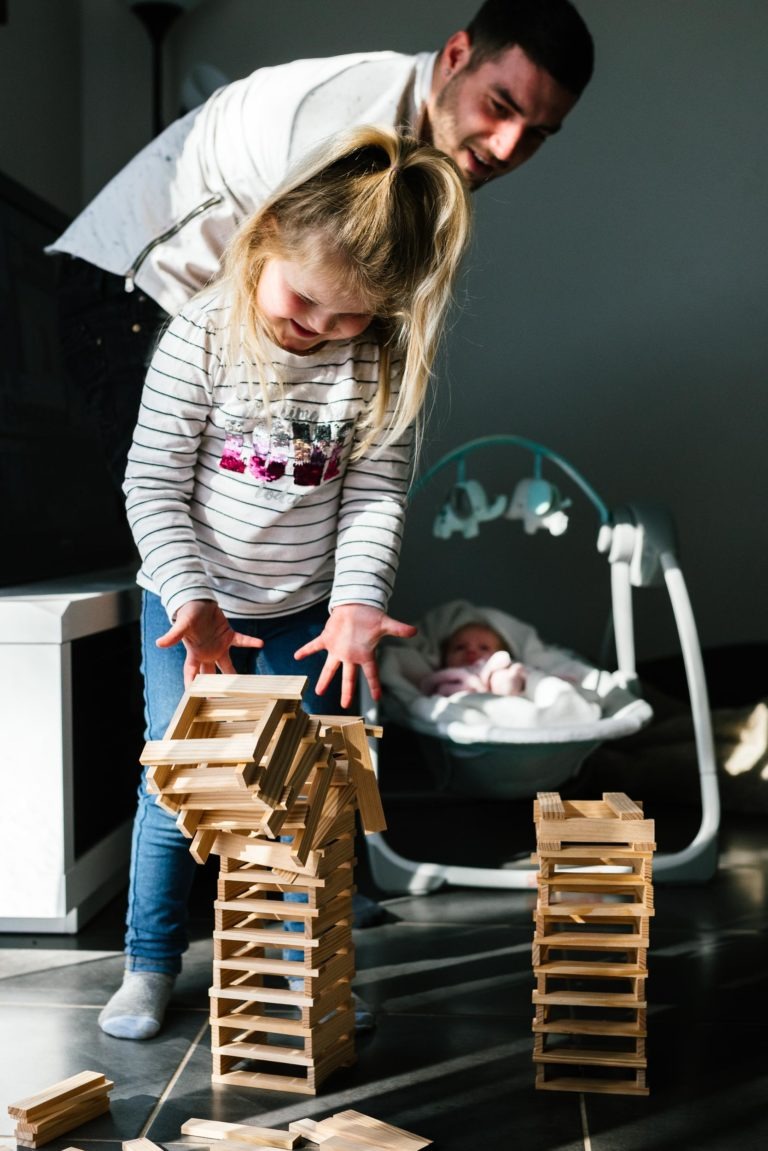 Une jeune fille aux cheveux blonds, vêtue d'une chemise rayée et d'un jean, construit avec enthousiasme une tour en bois. Un homme se tient derrière elle, souriant chaleureusement. En arrière-plan, un bébé se repose dans un transat. La scène chaleureusement éclairée ressemble à un coup de foudre au feu rouge, car la fille est profondément concentrée sur sa tâche.