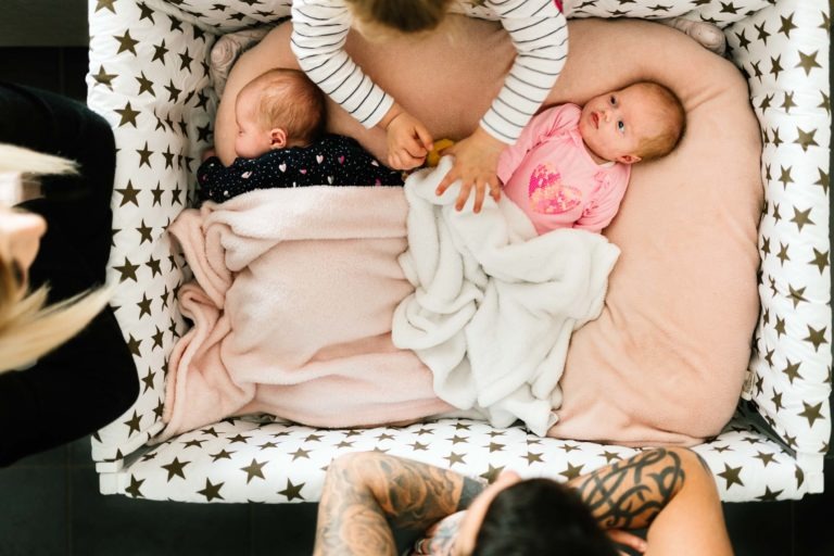 Deux bébés sont couchés dans un berceau dont la literie est décorée d'étoiles. Le bébé de gauche est emmailloté dans une couverture rose, et celui de droite est partiellement recouvert d'une couverture blanche, vêtu d'une tenue rose. Deux adultes sont visibles d'en haut, l'un avec des tatouages, regardant les bébés comme s'il s'agissait d'un coup de foudre au feu rouge, tandis que les mains d'un enfant touchent les couvertures.