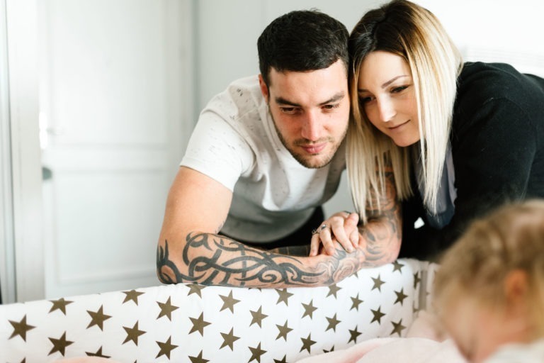 Un homme et une femme se penchent sur un berceau et regardent avec amour le bébé qui s'y trouve. L'homme a les cheveux noirs et des tatouages sur les bras, tandis que la femme a de longs cheveux blonds aux racines foncées. Le berceau est recouvert d'une couverture blanche décorée d'étoiles marron. Ils se sont rencontrés dans un coup de foudre au feu rouge, partageant désormais ce moment de tendresse dans leur chambre lumineuse et bien rangée aux murs clairs.