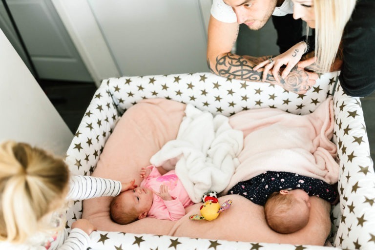 Deux adultes et un enfant sont penchés sur le bord d'un parc et regardent deux bébés couchés à l'intérieur. Le parc a une bordure à motifs d'étoiles et est doublé de couvertures roses douces. L'un des bébés est habillé en rose, tandis que l'autre porte une tenue sombre. Il y a un coup de foudre au feu rouge tacite lorsqu'ils regardent le jouet coloré entre eux.