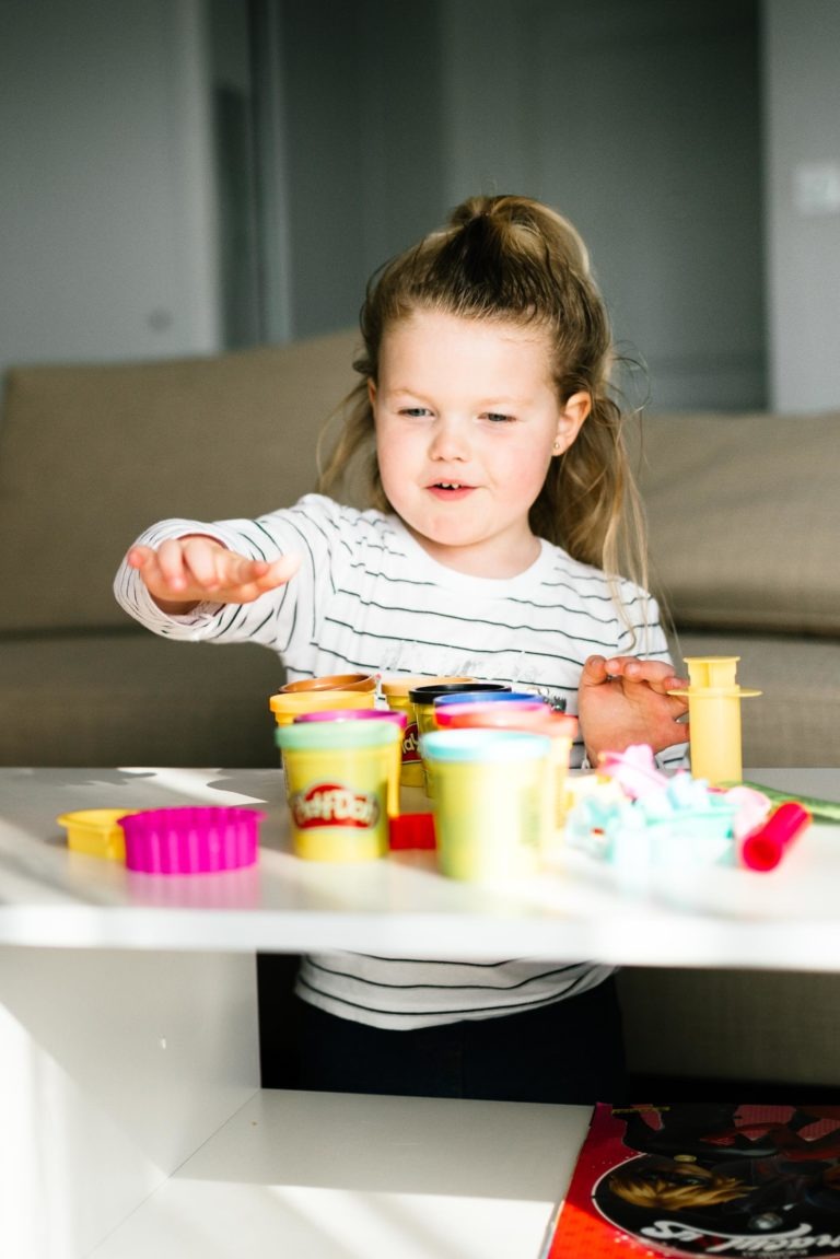 Une jeune fille aux cheveux longs attachés en deux joue avec de la pâte à modeler colorée Play-Doh, rappelant un coup de foudre au feu rouge. Elle est assise à une table, tendant la main vers divers contenants et moules Play-Doh étalés devant elle. Elle porte une chemise blanche à manches longues à rayures noires, tandis qu'un canapé beige trône à l'arrière-plan.